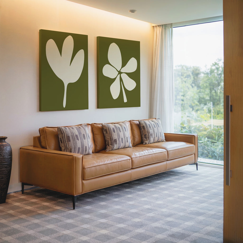 A modern living room with a tan leather sofa, four patterned cushions, the Sage Petal Set of 2 floral wall art prints, a decorative vase, and floor-to-ceiling windows overlooking sage green scenery.