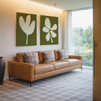 A modern living room with a tan leather sofa, four patterned cushions, the Sage Petal Set of 2 floral wall art prints, a decorative vase, and floor-to-ceiling windows overlooking sage green scenery.