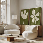 A modern living room features two white rounded armchairs, a low wooden coffee table, a floor lamp, and the Sage Petal Set of 2 floral wall art prints on the console. Sunlight streams through tall windows.