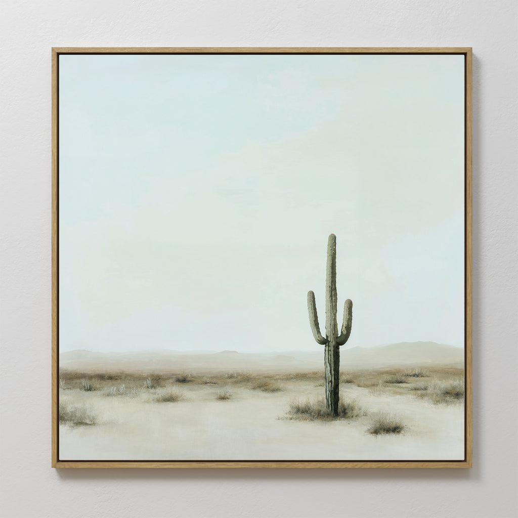 The Saguaro Whisper Canvas Art is a large framed painting showcasing a minimalist desert landscape with a central tall saguaro cactus, sparse dry grass, and a soft blue sky with light clouds.