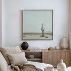 A minimal, modern living room features a beige sofa, neutral cushions, wooden sideboard, vases, books, and a large Saguaro Whisper Canvas Art—framed desert landscape with a lone saguaro cactus—displayed on the wall.