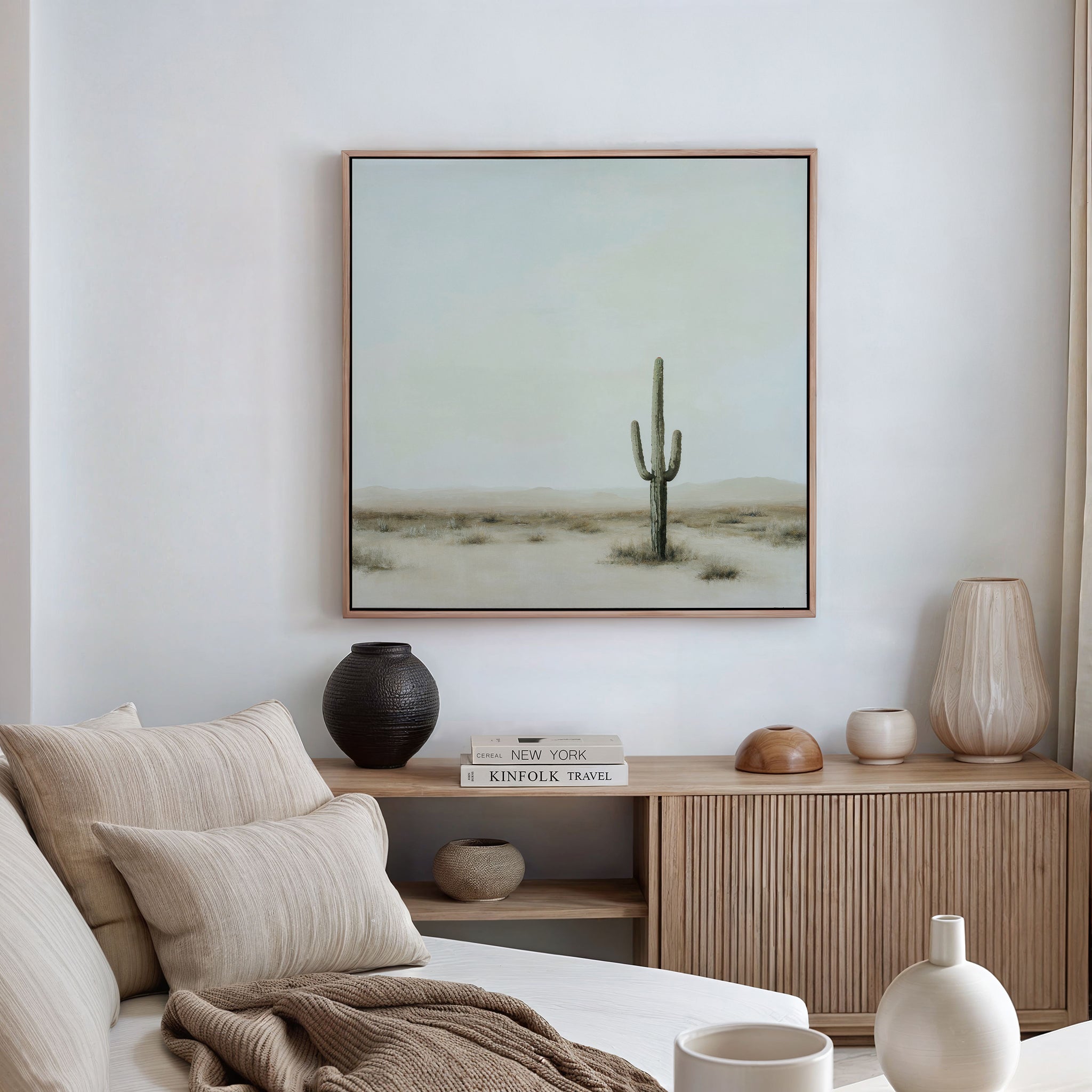 A minimal, modern living room features a beige sofa, neutral cushions, wooden sideboard, vases, books, and a large Saguaro Whisper Canvas Art—framed desert landscape with a lone saguaro cactus—displayed on the wall.