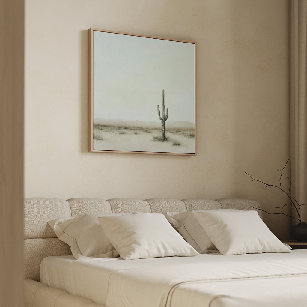 A minimalist bedroom features a beige upholstered bed with four white pillows and the Saguaro Whisper Canvas Art hanging above the headboard. Neutral tones define the room’s decor.