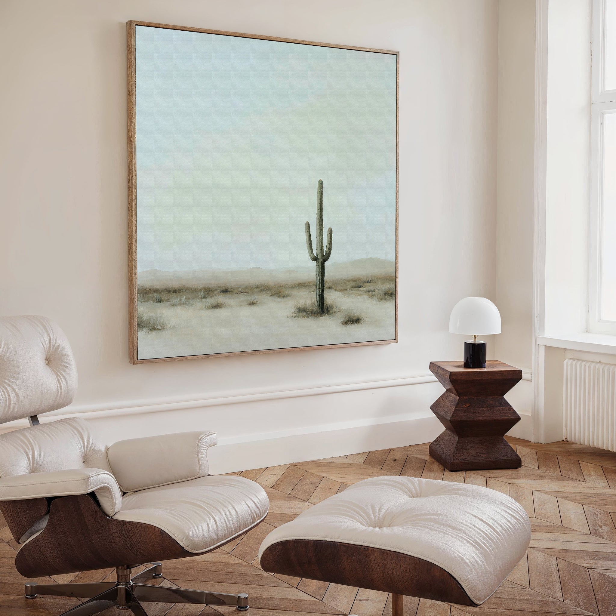 A modern, minimal living room with a white leather lounge chair and ottoman, wooden side table with a lamp, large window, and the Saguaro Whisper Canvas Art—a framed minimalist desert landscape with a saguaro cactus—on the wall.