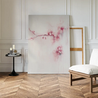 The Sakura Dream Canvas Art featuring soft pink cherry blossoms leans against a wall beside an empty frame. A round black side table with candles and books stands next to a white cushioned chair on a herringbone wood floor.