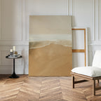 The Sand and Sea Abstract Canvas Art, depicting a serene beach with gentle waves, leans against a white-paneled wall beside an empty wooden frame, a modern cushioned chair, and a round table—ideal for neutral home decor.