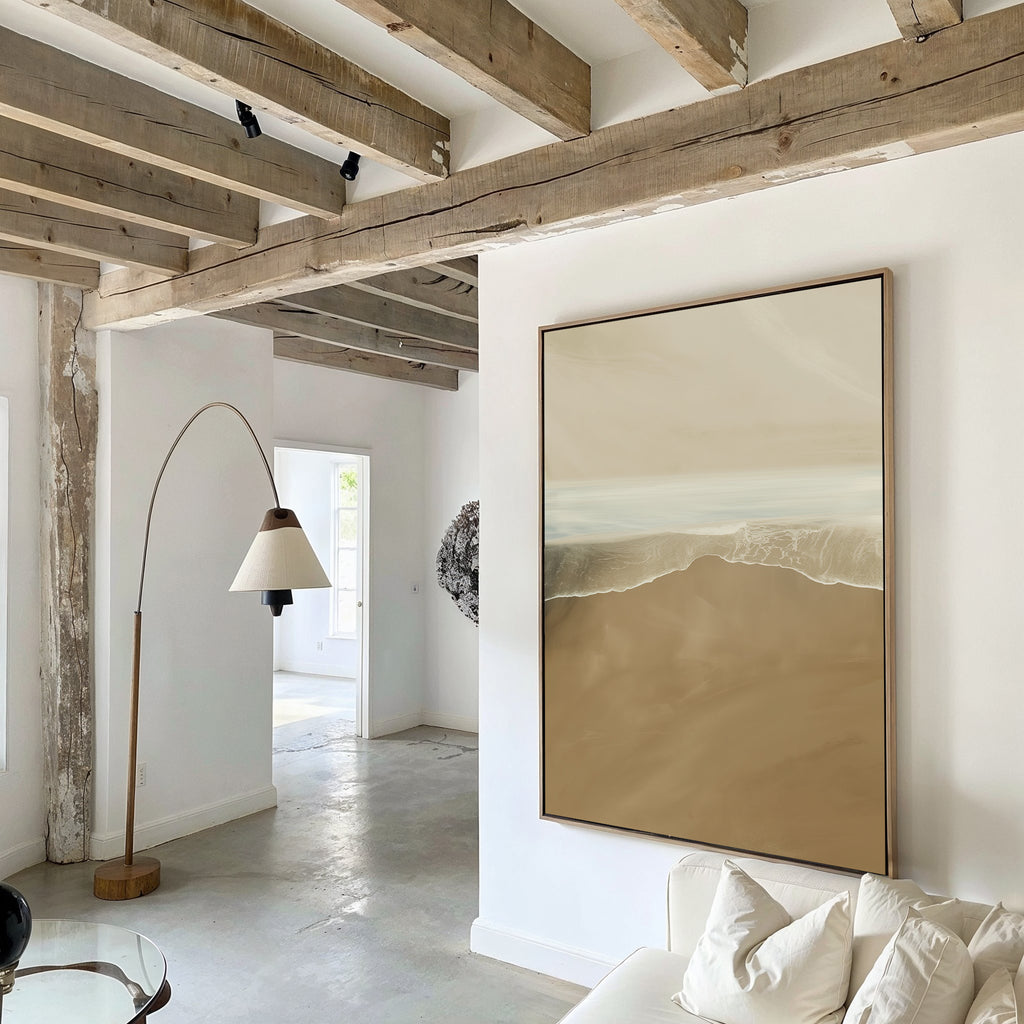 A modern living room with exposed wooden ceiling beams, white walls, Sand and Sea Abstract Canvas Art above the sofa, a curved floor lamp, neutral decor, and natural light filling the space.