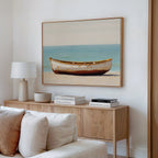 A minimalist living room features a beige couch, a wooden sideboard with books and decor, and the Sandbound Vessel Canvas Art—a striking coastal wall piece depicting a weathered boat on a sandy beach with blue water behind.