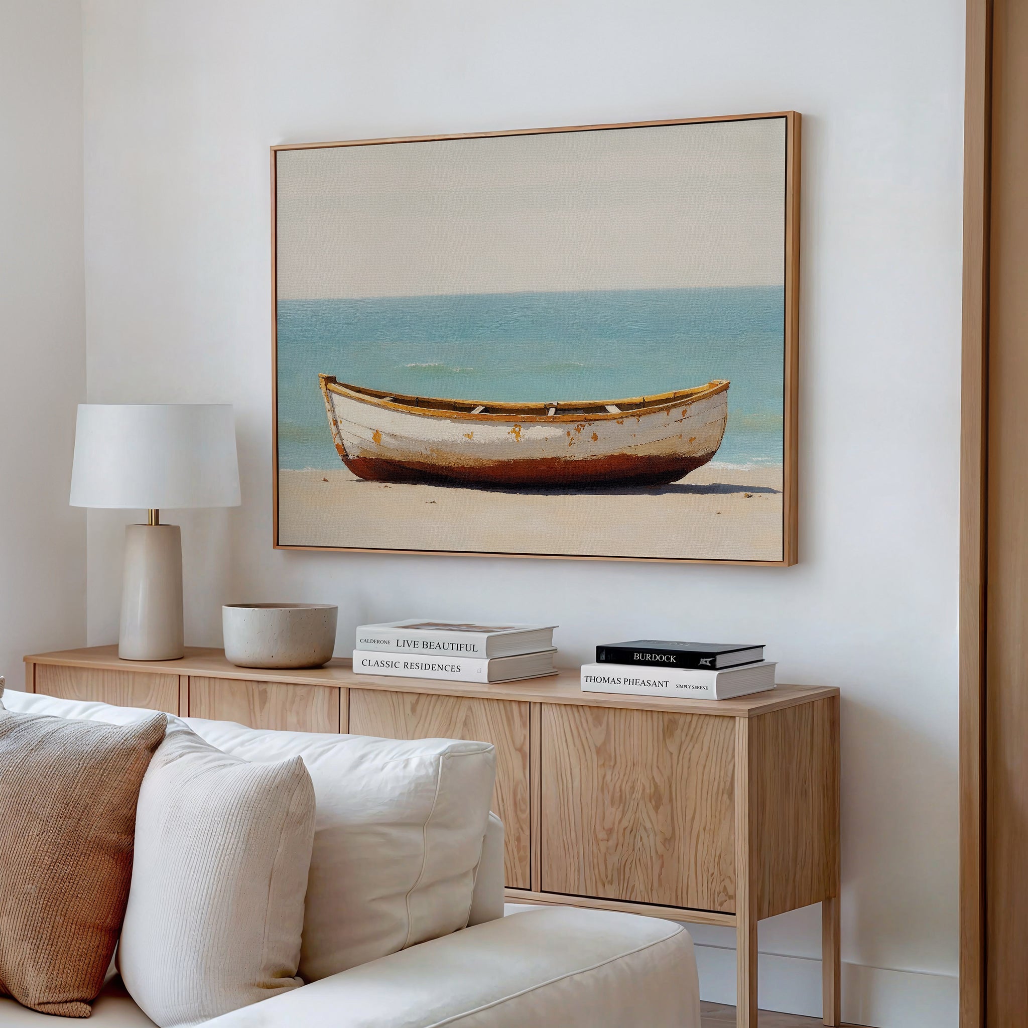 A minimalist living room features a beige couch, a wooden sideboard with books and decor, and the Sandbound Vessel Canvas Art—a striking coastal wall piece depicting a weathered boat on a sandy beach with blue water behind.