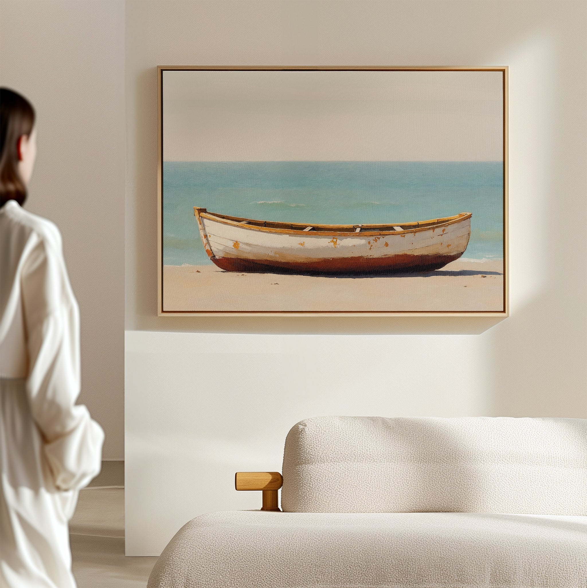 A person in white admires the Sandbound Vessel Canvas Art—a framed painting of an old white boat on a sandy beach—hanging above a light sofa in a bright, minimalist room. Ideal for adding serenity to bedroom decor.