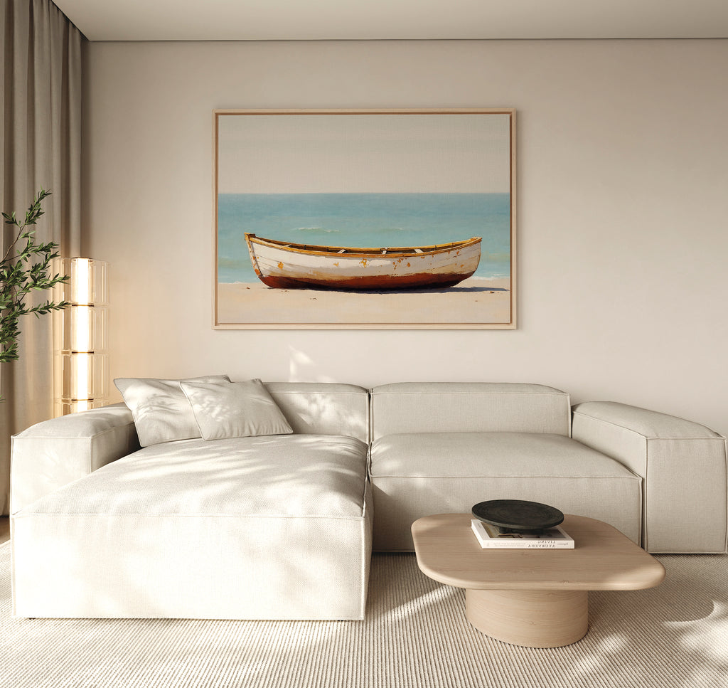 A modern living room with a light beige sectional sofa, a low wooden coffee table, and the Sandbound Vessel Canvas Art—featuring a boat on the beach—framed above the sofa. A plant stands in the corner by the window.