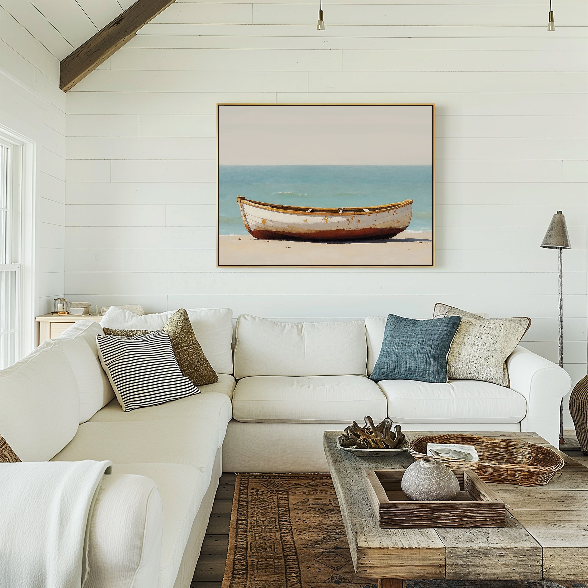 A cozy living room featuring a white sectional sofa, neutral pillows, woven accents, and coastal charm with the Sandbound Vessel Canvas Art—showing a boat on the beach—displayed on a white shiplap wall.