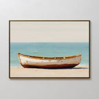The Sandbound Vessel Canvas Art depicts a weathered wooden rowboat on a sandy shore with calm blue water and a pale sky, creating striking coastal wall art ideal for serene bedroom decor.