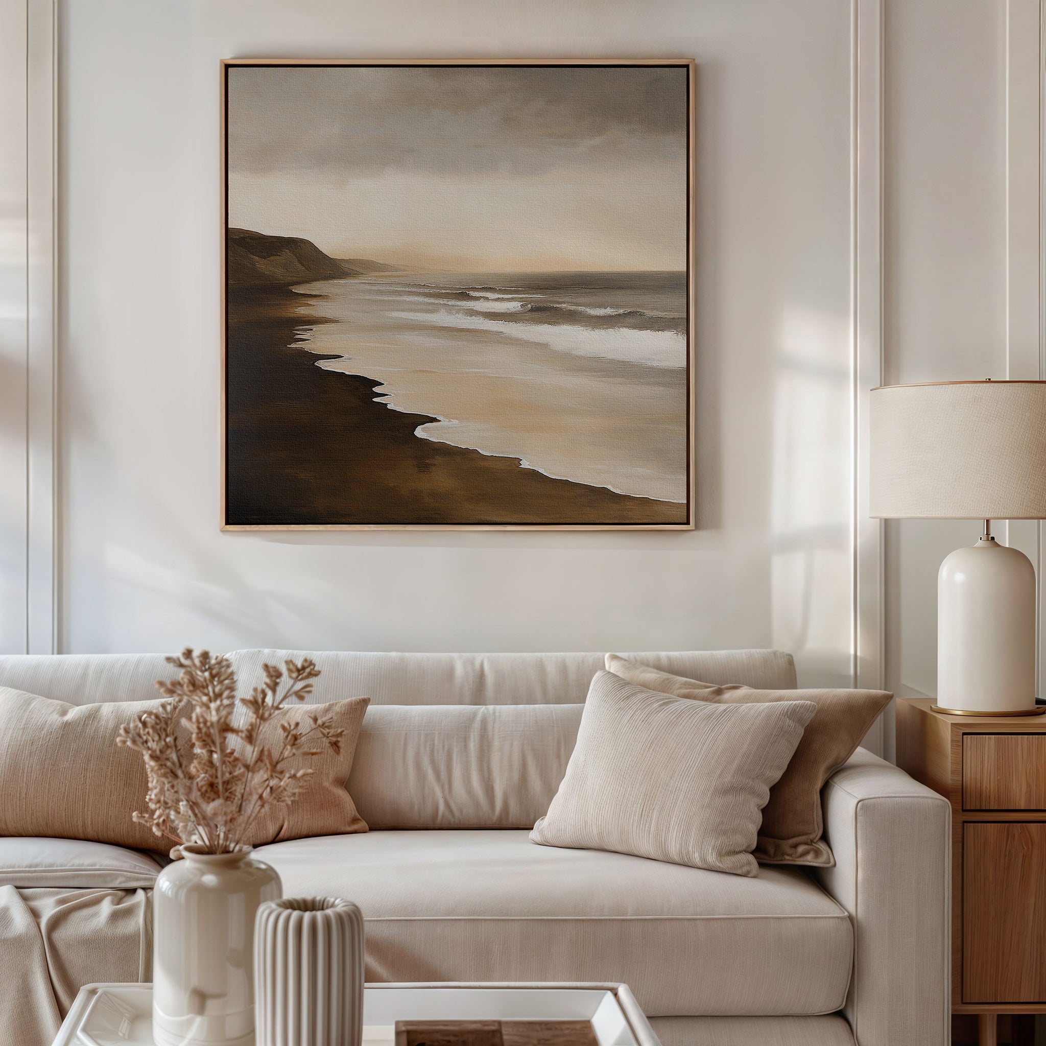A neutral-toned living room with a beige sofa, cream pillows, a table lamp, and a vase with dried flowers features the Sandy Shoreline Canvas Art—a large coastal beach scene—hanging above the sofa.