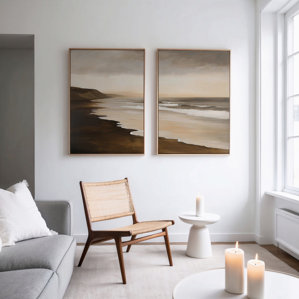 A modern living room with a wooden chair, small white table, lit candles, gray sofa with a white pillow, and the Sandy Shoreline Set of 2 beach canvases hanging above. Natural light pours in from tall windows.