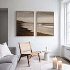 A modern living room with a wooden chair, small white table, lit candles, gray sofa with a white pillow, and the Sandy Shoreline Set of 2 beach canvases hanging above. Natural light pours in from tall windows.