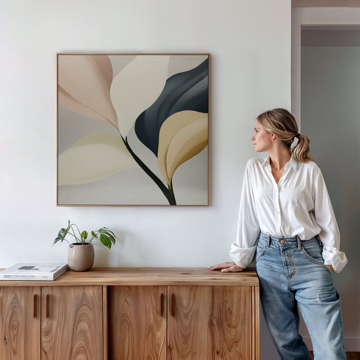 A woman in a white blouse and jeans leans against a wooden cabinet, admiring the Sculpted Elegance Canvas Art. A small plant and books rest on the cabinet, adding finishing touches to this minimalistic, bright living room decor scene.