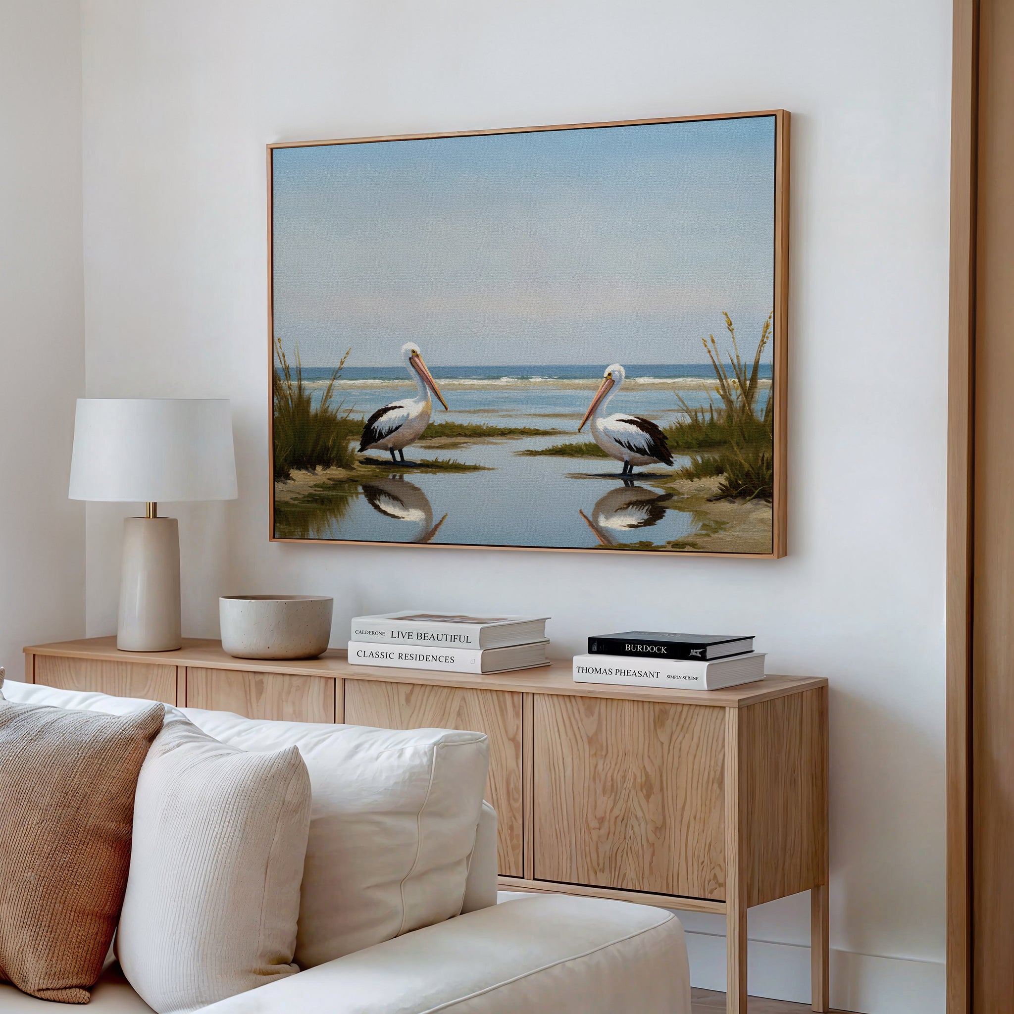 A modern living room features a wooden console with books, lamp, and decorative bowls, topped by the Seaside Gaze Canvas Art depicting two pelicans by the sea. A white sofa is partially visible in the foreground.