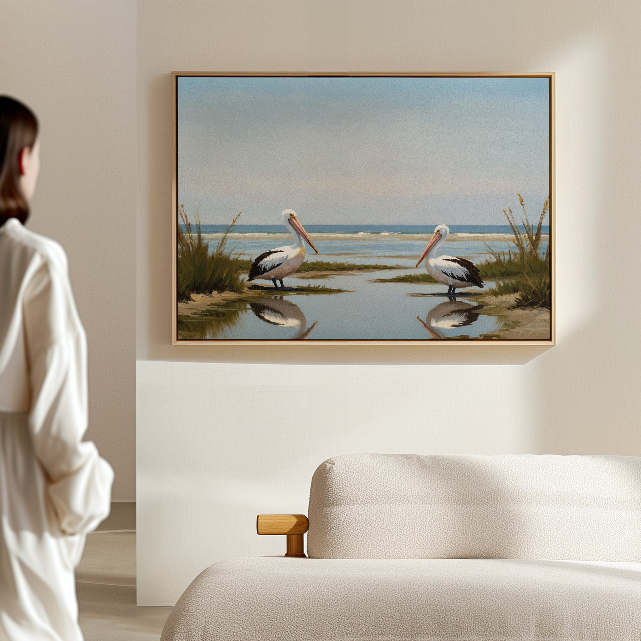 In a minimalist room, a person admires the Seaside Gaze Canvas Art—a framed painting of two pelicans by the water with reeds and ocean—displayed above a light-colored sofa.