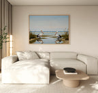 A modern living room features a light beige sectional, a round wooden coffee table, and the Seaside Gaze Canvas Art—a beach scene with two pelicans—framed above the sofa. Sunlight filters through large windows dressed in sheer curtains.