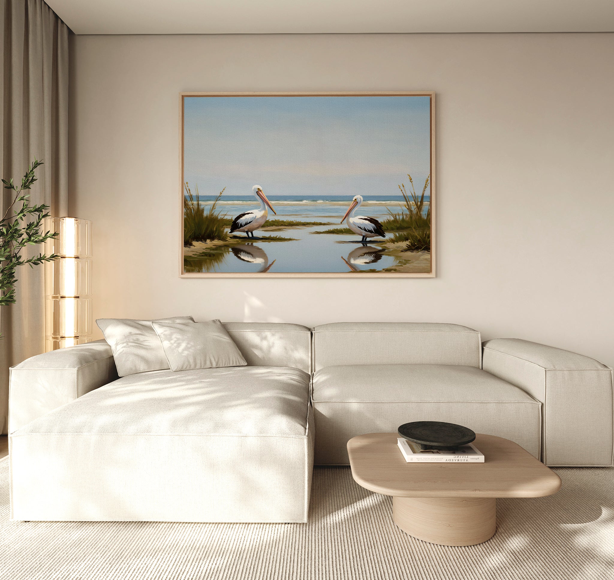 A modern living room features a light beige sectional, a round wooden coffee table, and the Seaside Gaze Canvas Art—a beach scene with two pelicans—framed above the sofa. Sunlight filters through large windows dressed in sheer curtains.