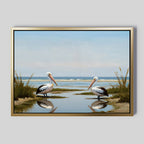 The Seaside Gaze Canvas Art features two pelicans by a calm lagoon with tall grass, blue sky, and ocean waves, their reflections shimmering in the water—adding a touch of serene coastal beauty to any space.