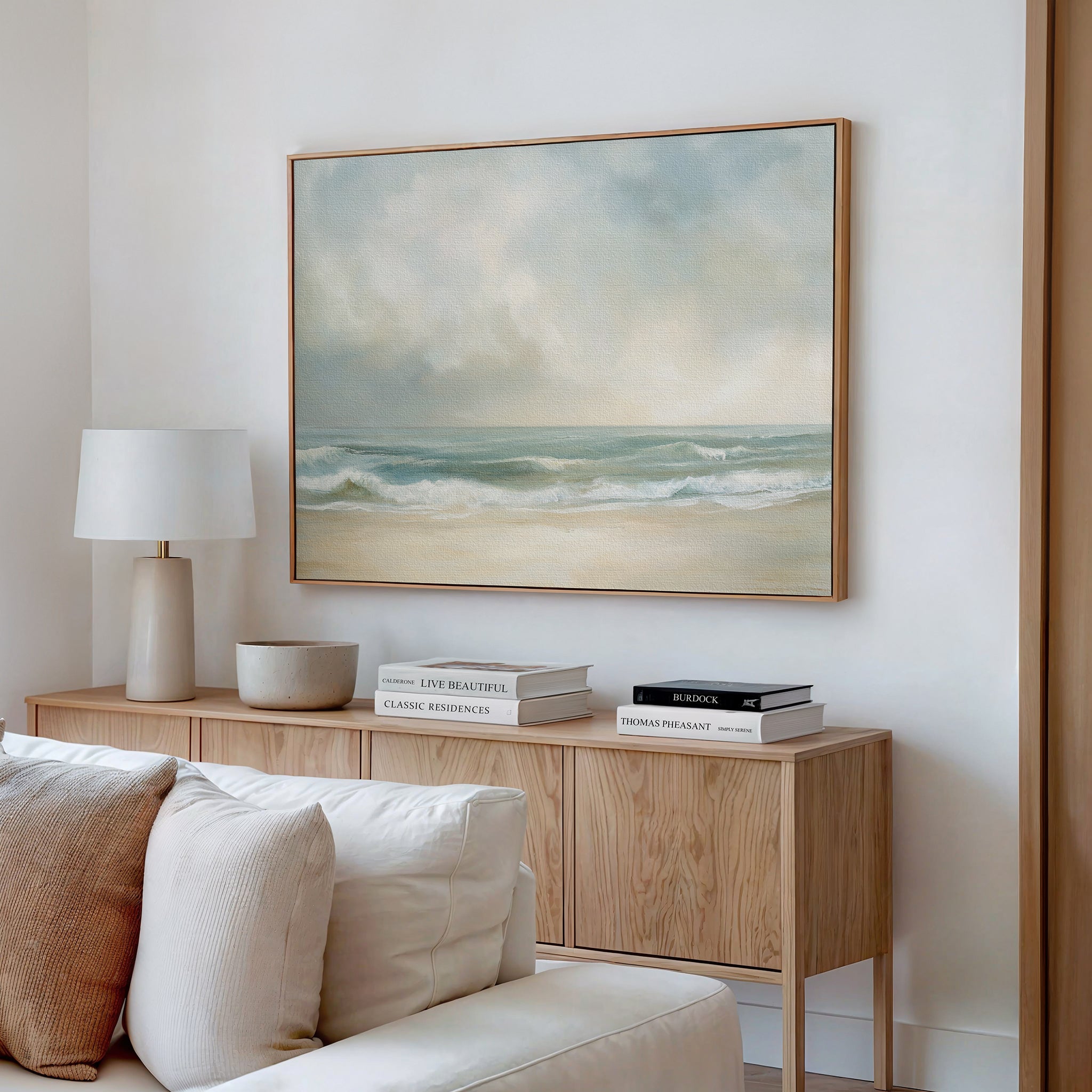 A modern living room with a wooden sideboard, stacked books, a white lamp, and neutral décor. Above the sideboard, a large framed Serene Beach Canvas Art adds calming vibes to the space.