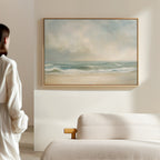 A person in a white shirt admires the Serene Beach Canvas Art featuring soft waves and a cloudy sky, hanging above a light sofa in a bright, minimalist room and adding vintage charm to the decor.