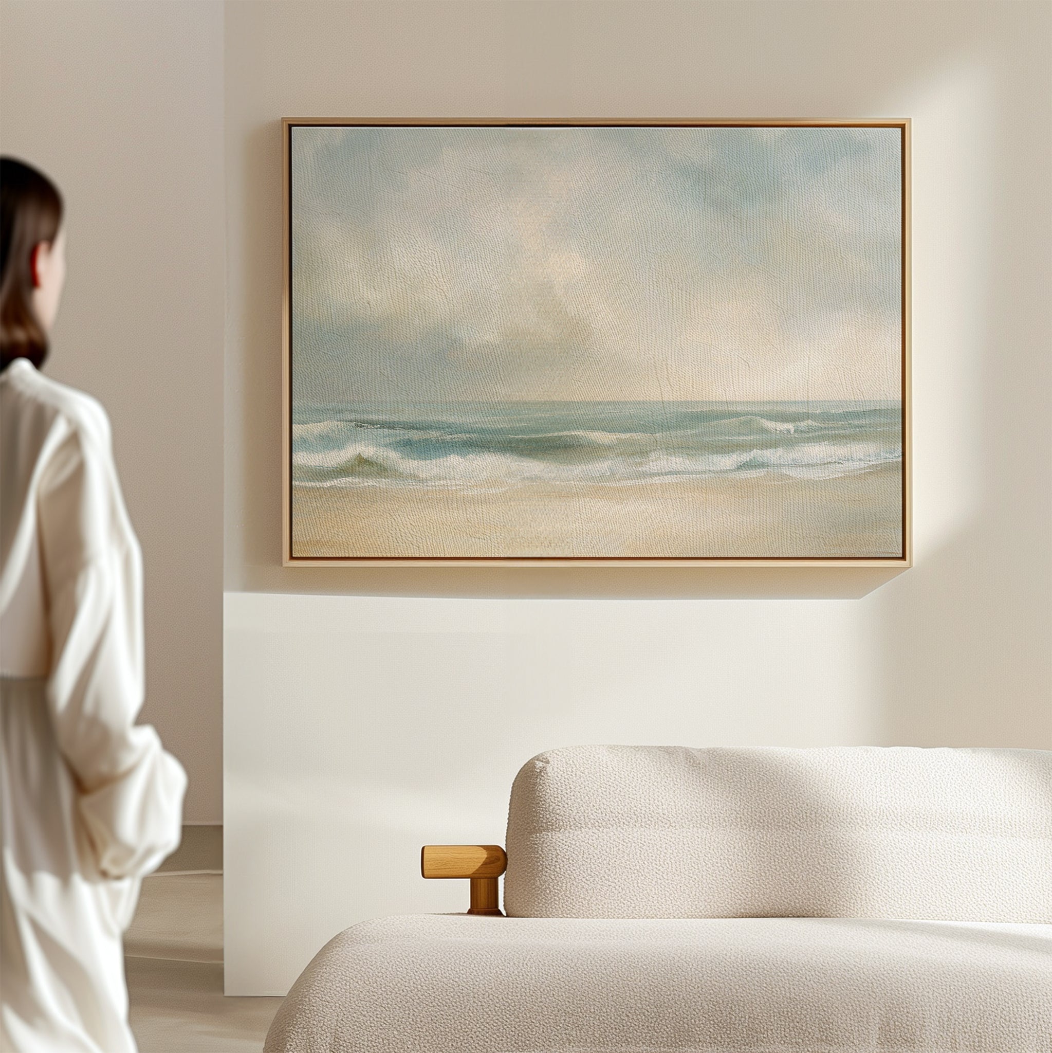 A person in a white shirt admires the Serene Beach Canvas Art featuring soft waves and a cloudy sky, hanging above a light sofa in a bright, minimalist room and adding vintage charm to the decor.