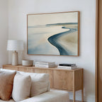 A minimalist living room with a wooden sideboard, stacked books, a white lamp, and the Serene Coastline Canvas Art – Horizontal—a framed abstract beach scene in soft blue and beige—adds a modern, coastal touch to the white wall decor.