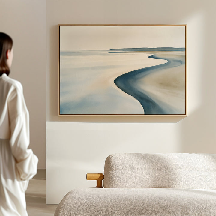 A woman admires the Serene Coastline Canvas Art – Horizontal, a framed abstract landscape in soft blue and beige tones, displayed above a light sofa in a minimalist room, enhancing the modern home decor.