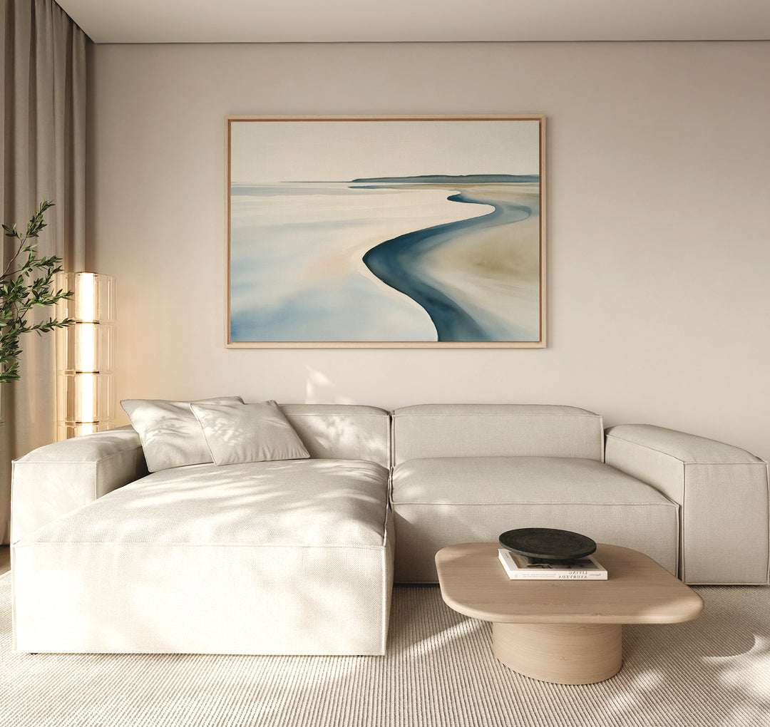 A modern living room features a light beige sectional sofa, low wooden coffee table, and the Serene Coastline Canvas Art – Horizontal, whose soft blue and sandy hues add to the calm, inviting atmosphere with minimalist neutral decor.