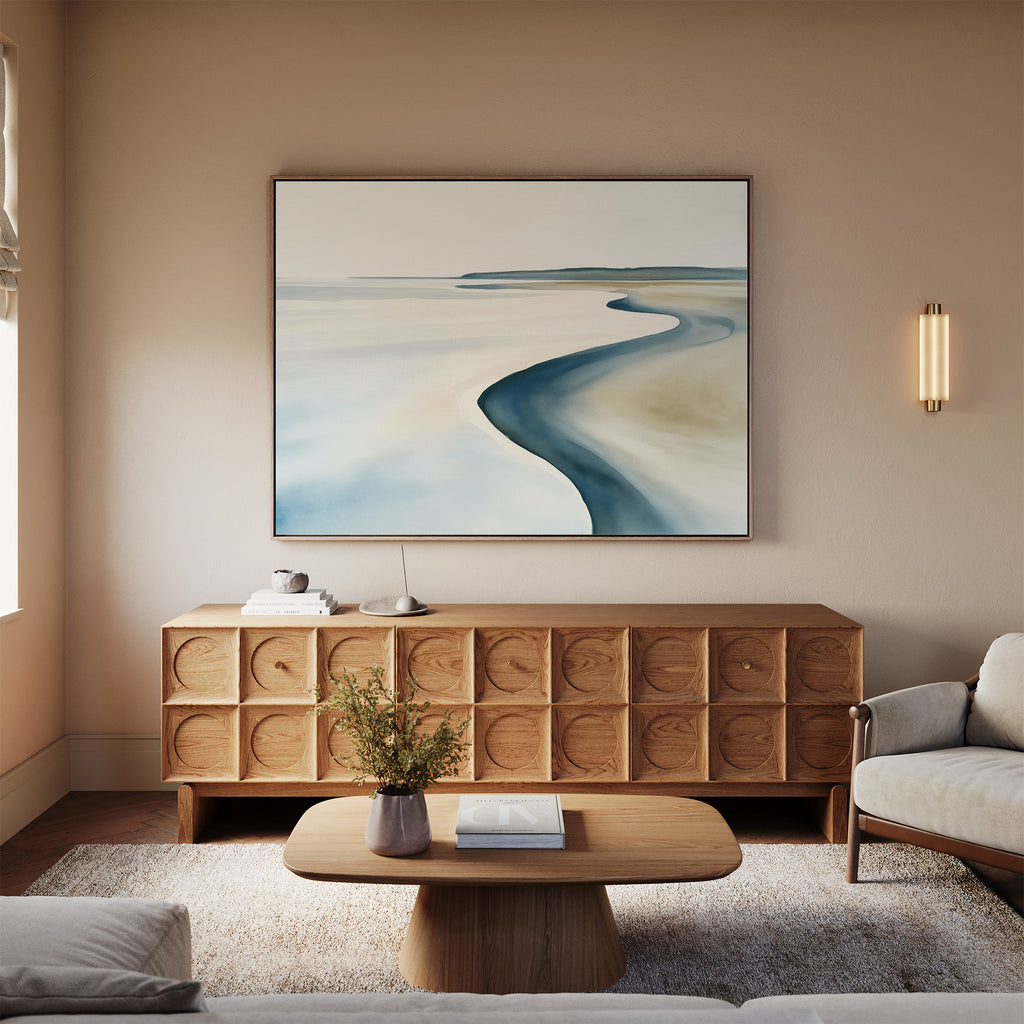A minimalist living room features a light wood credenza, beige walls with the Serene Coastline Canvas Art – Horizontal above, a round coffee table with a small flower vase, a neutral sofa, and a modern wall sconce illuminating the space.