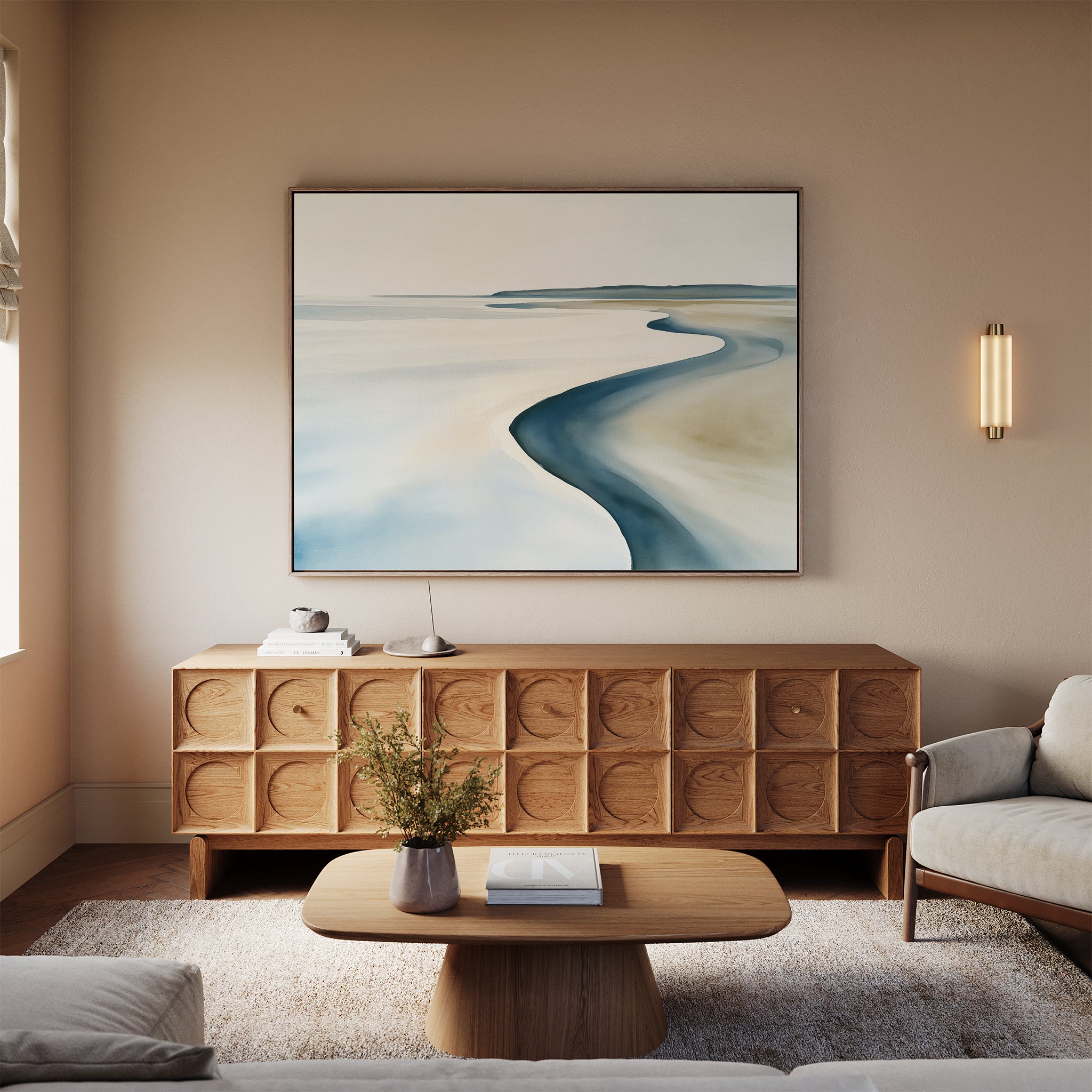 A minimalist living room features a light wood credenza, beige walls with the Serene Coastline Canvas Art – Horizontal above, a round coffee table with a small flower vase, a neutral sofa, and a modern wall sconce illuminating the space.