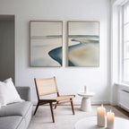 A minimalist living room with a wicker chair, small white table, lit candles, gray sofa, and the Serene Coastline Set of 2 wall art hanging on a white wall near a large window.
