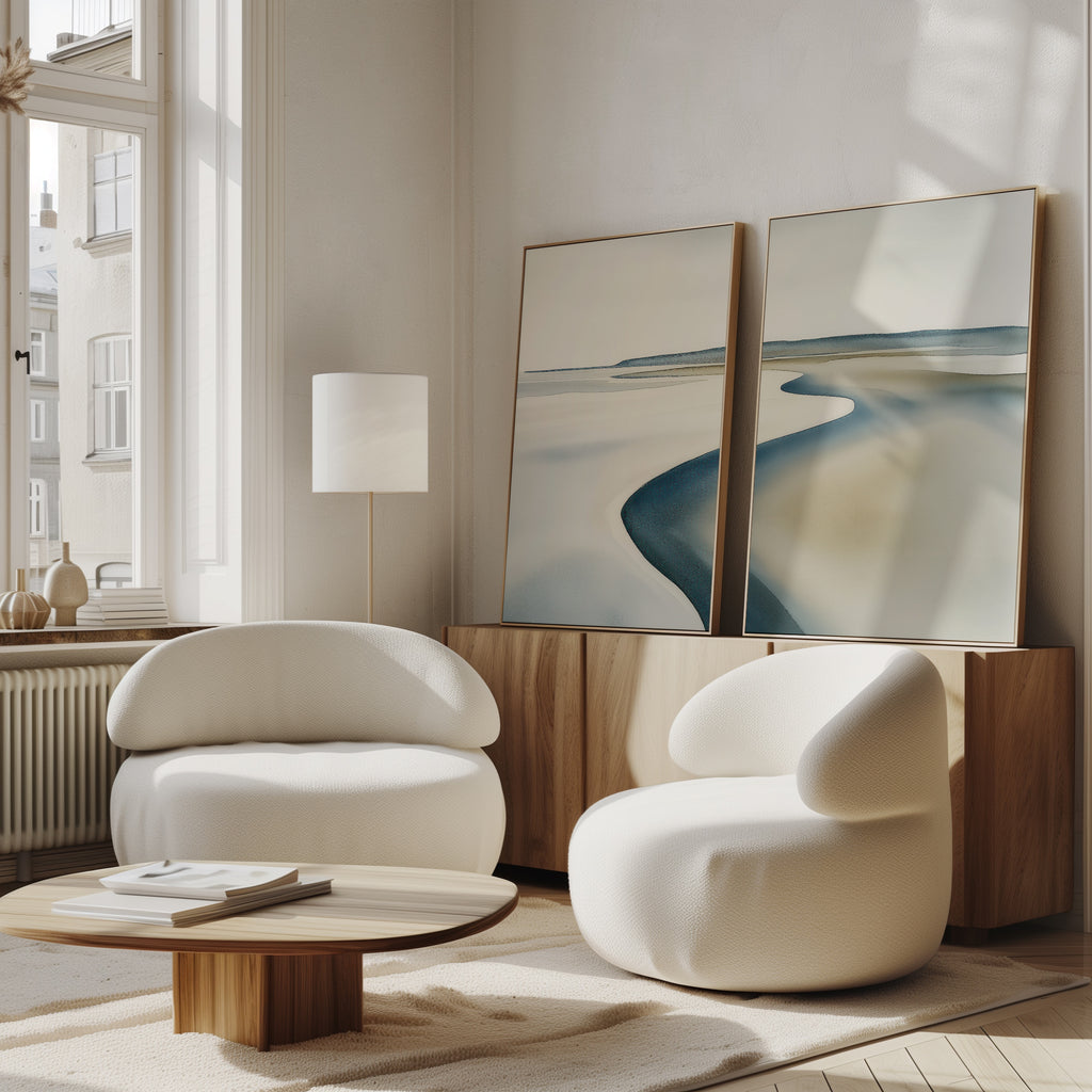 A modern living room with the Serene Coastline Set of 2 curved white armchairs, a low wooden coffee table, a sideboard, and large minimalist wall art leaning under a tall window with soft natural light.