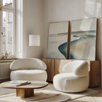 A modern living room with the Serene Coastline Set of 2 curved white armchairs, a low wooden coffee table, a sideboard, and large minimalist wall art leaning under a tall window with soft natural light.