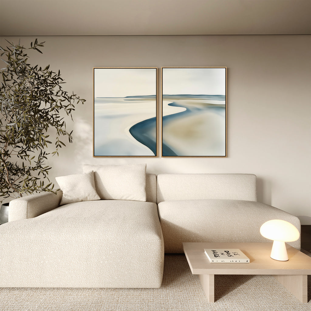 A modern living room with a cream sectional sofa, a wooden coffee table holding a small round lamp and book, an indoor plant, and the Serene Coastline Set of 2 minimalist wall art on a beige wall.