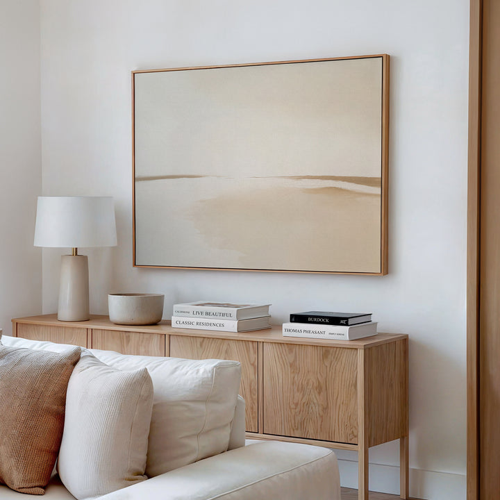 A minimalist living room features a light wood sideboard with books, a white lamp, and a bowl. Above, the Serene Expanse Canvas Art – Horizontal in beige tones decorates the white wall. A cream sofa is partially visible in the modern space.