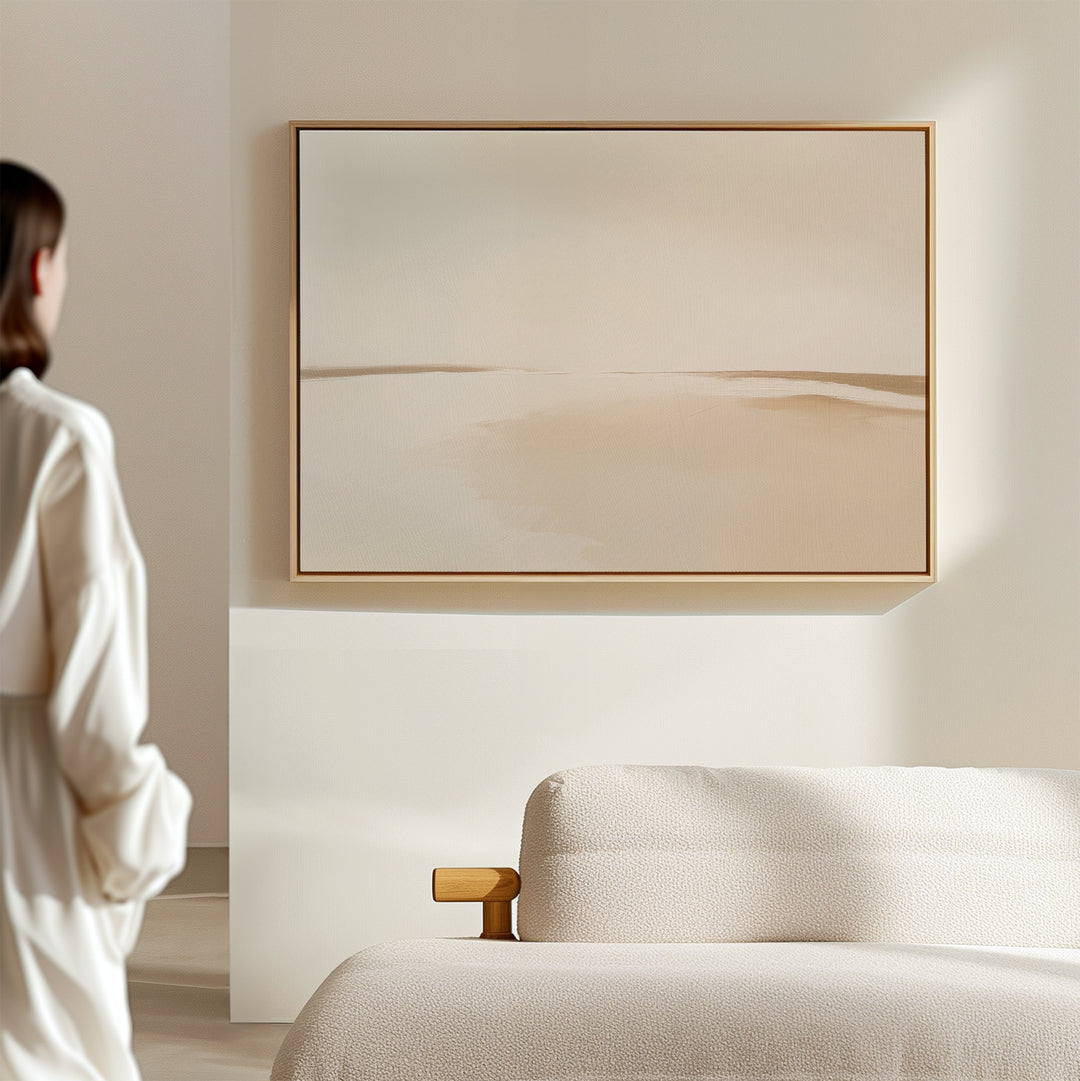 A woman in a cream outfit stands in a modern space with a light sofa and the Serene Expanse Canvas Art – Horizontal, an abstract landscape in soft beige tones, adding elegance to the living room decor.