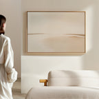 A woman in a cream outfit stands in a modern space with a light sofa and the Serene Expanse Canvas Art – Horizontal, an abstract landscape in soft beige tones, adding elegance to the living room decor.