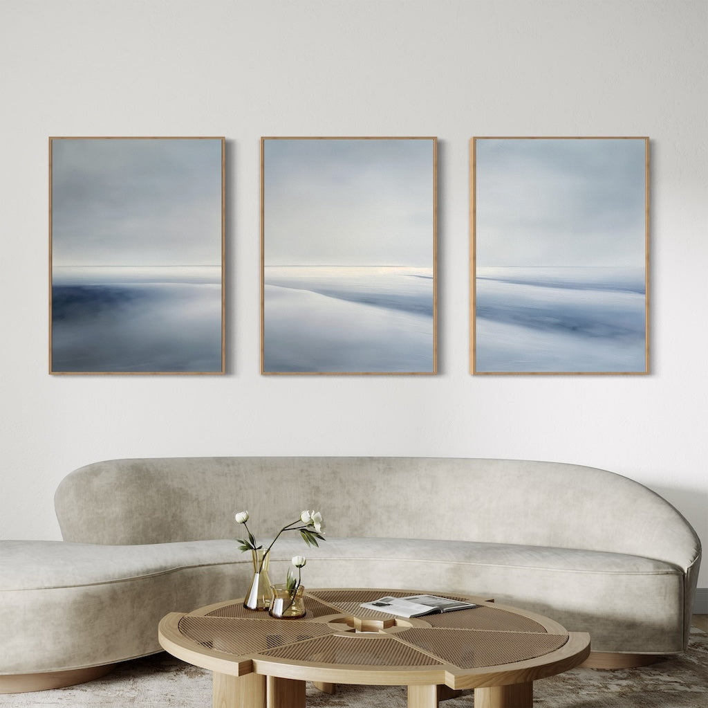 A modern living room with a curved beige velvet sofa, a round wooden coffee table topped with a glass vase of white flowers, and the Serene Horizons Set of 3 abstract landscape paintings in soft blue and gray tones creates an inviting atmosphere.