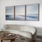 A modern living room features a curved cream sofa, a round wooden coffee table with a vase and book, and the Serene Horizons Set of 3 abstract landscape artwork in soft blue and gray.