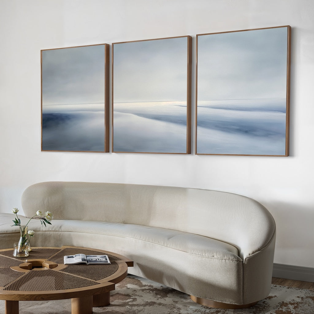 A modern living room features a curved cream sofa, a round wooden coffee table with a vase and book, and the Serene Horizons Set of 3 abstract landscape artwork in soft blue and gray.