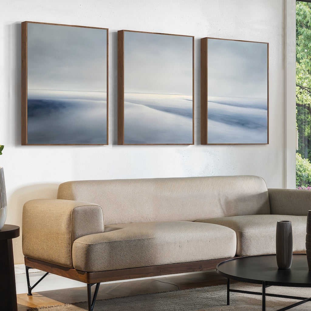 A modern living room features a beige sectional sofa, round black coffee table, and the Serene Horizons Set of 3—large abstract landscape paintings in wooden frames—on a white wall. A window showcases greenery outside.