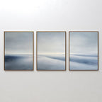 The Serene Horizons Set of 3 features framed abstract wall art prints with soft, blurred shades of blue, gray, and white—arranged side by side to create a calming, minimalist landscape on any wall.
