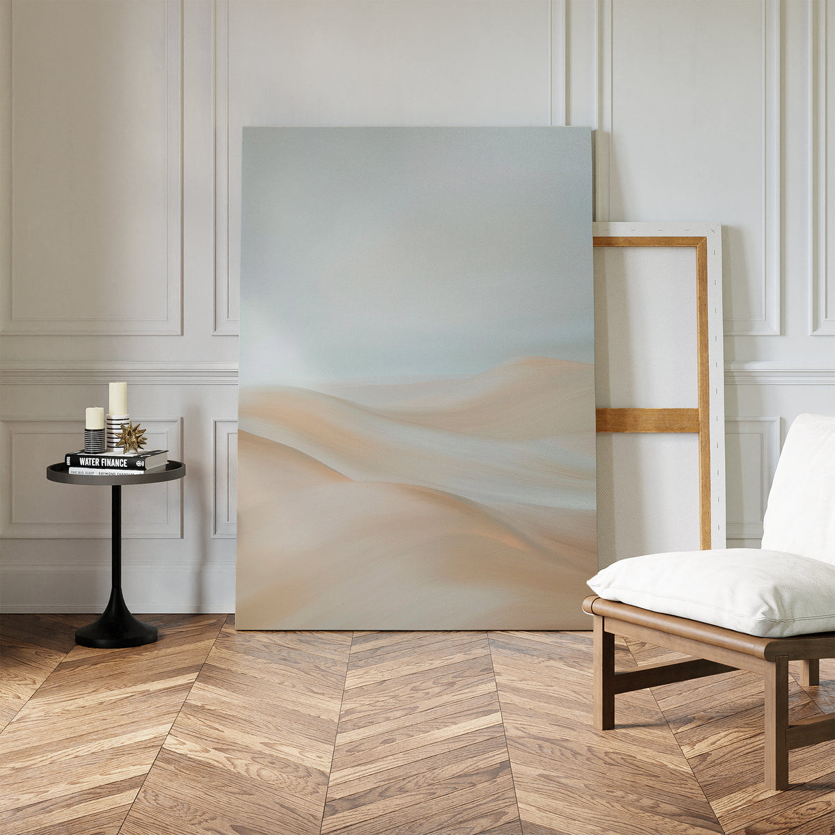 A minimalist room features herringbone wood floors, a white cushioned chair, a round black side table with candles and books, and the Serene Peaks Abstract Canvas Art leaning against a white paneled wall for calm elegance.