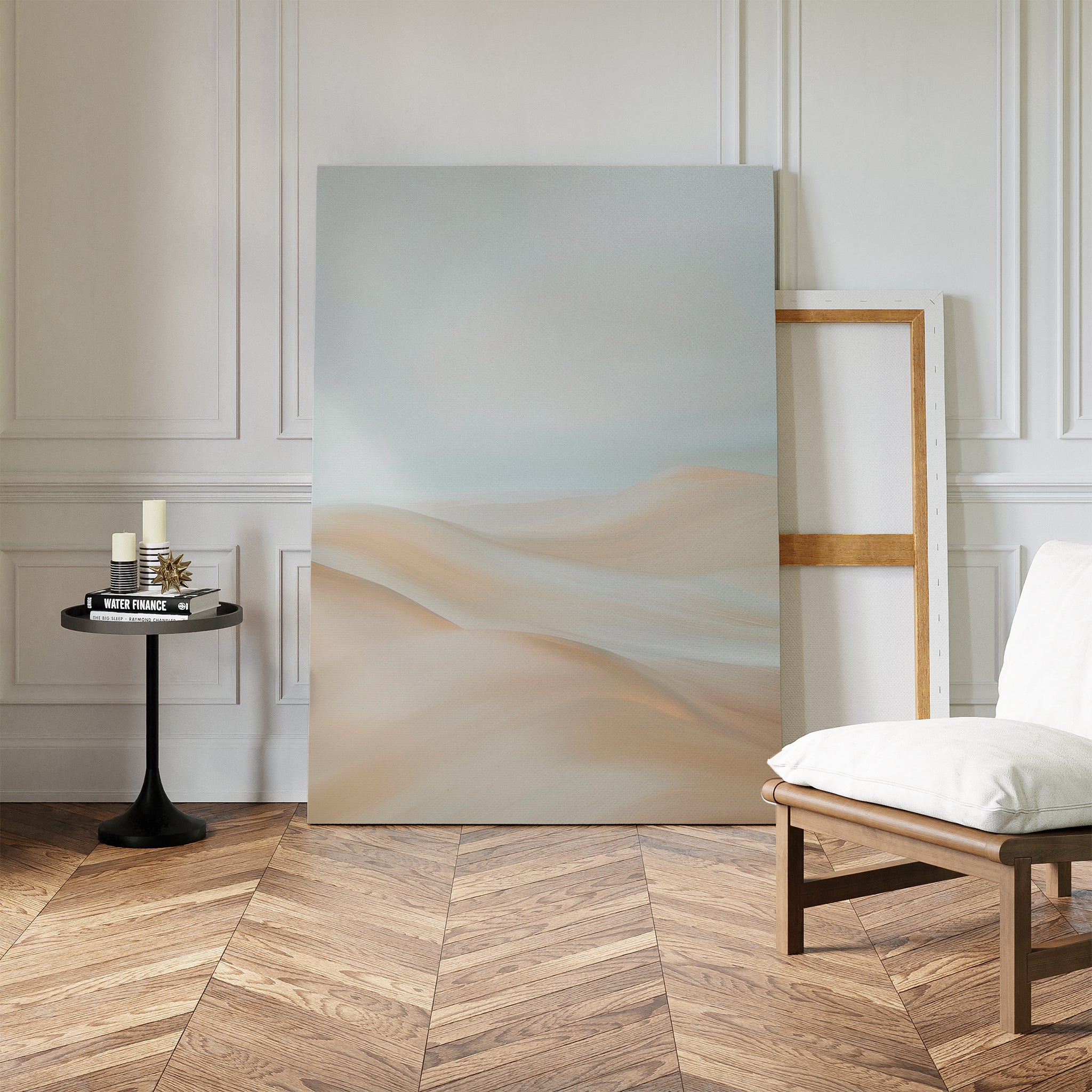 A minimalist room features herringbone wood floors, a white cushioned chair, a round black side table with candles and books, and the Serene Peaks Abstract Canvas Art leaning against a white paneled wall for calm elegance.