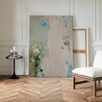 The Serenity Garden Abstract Canvas Art, featuring soft blue, green, and beige tones, rests against a white wall beside an empty wooden frame. Nearby are a black table with candles and books, a cushioned chair, and herringbone wood flooring.