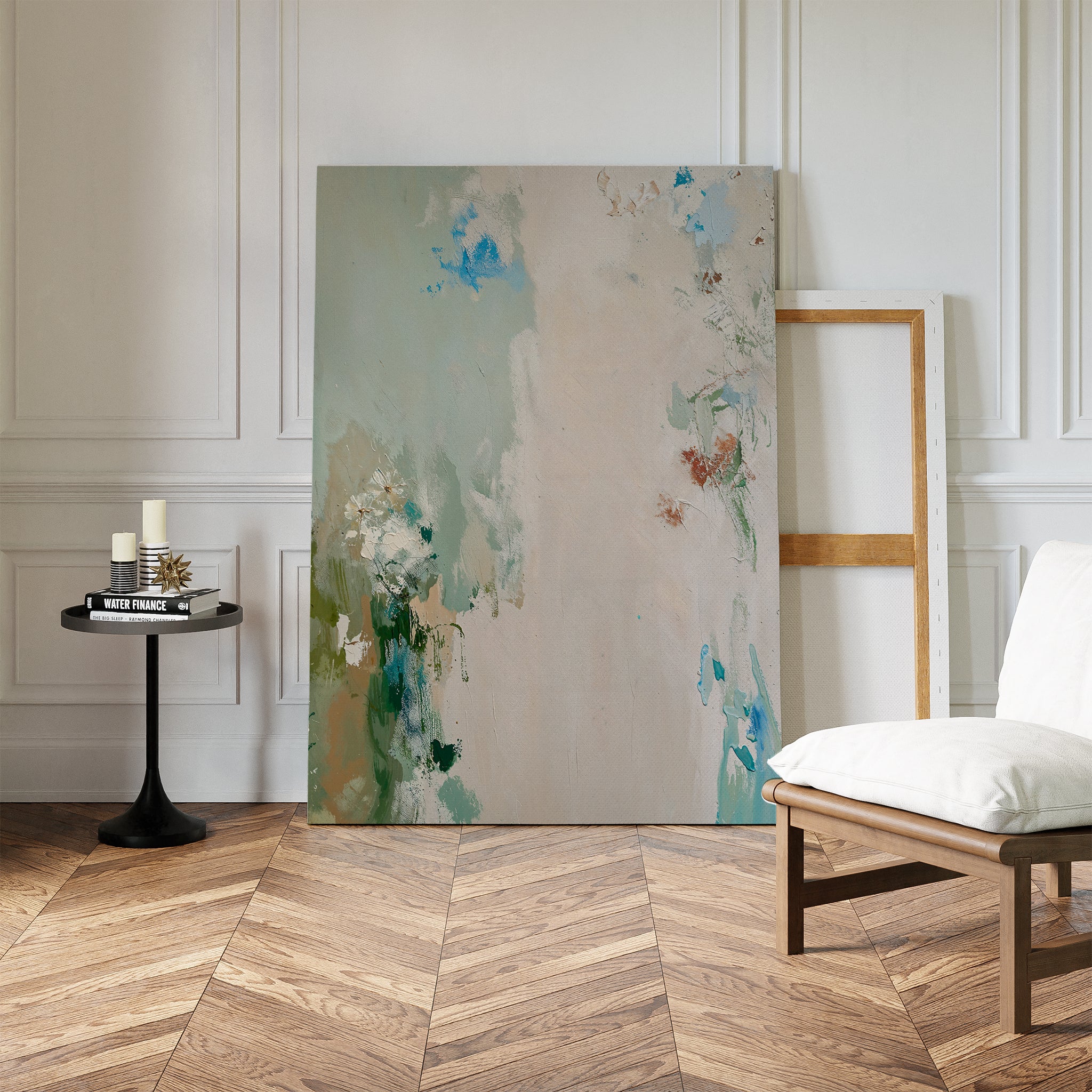 The Serenity Garden Abstract Canvas Art, featuring soft blue, green, and beige tones, rests against a white wall beside an empty wooden frame. Nearby are a black table with candles and books, a cushioned chair, and herringbone wood flooring.